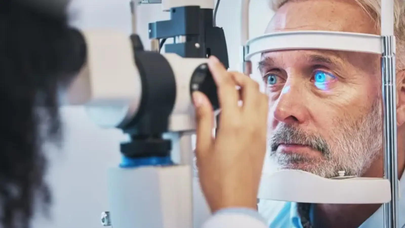 A man undergoing an eye examination. He is looking into a machine as a healthcare professional adjusts it. The machine's light is glowing on his eye, and he appears focused on the test.